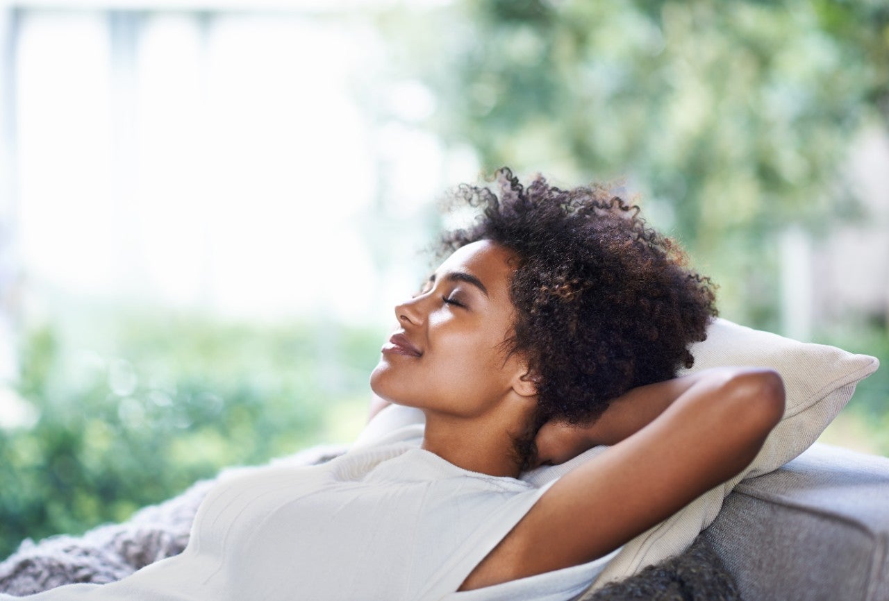 A woman relaxing on a couch with her hands behind her head, eyes closed and smiling peacefully, surrounded by soft natural light and a blurred green outdoor background.