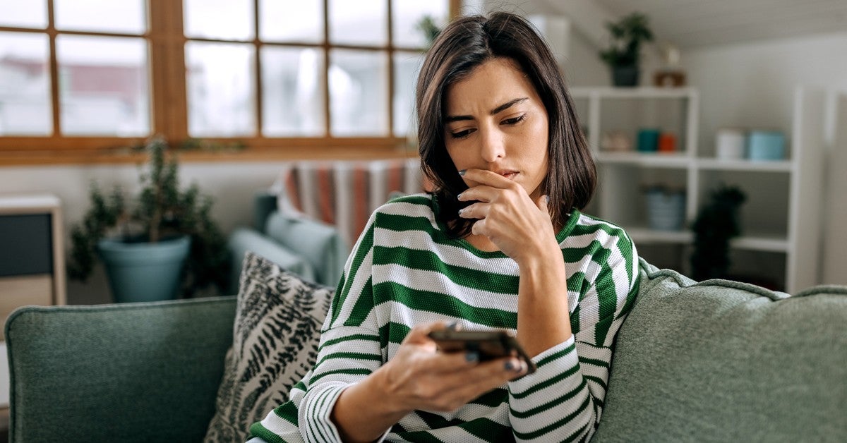 Young woman sitting on couch looking at her phone with a confused look