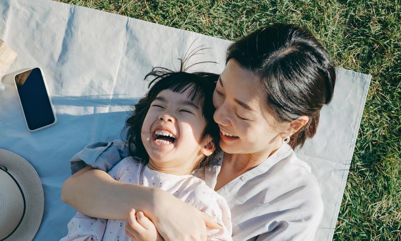 A mother and child lying on a picnic blanket, laughing and hugging together..
