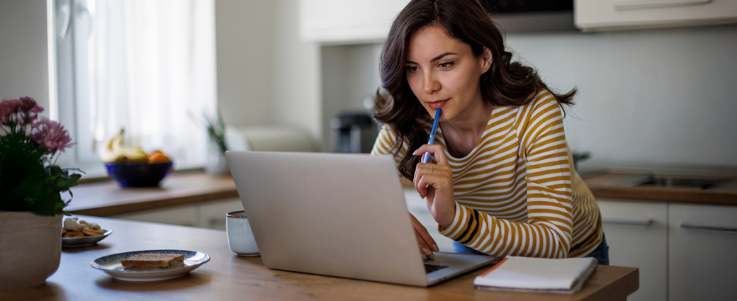 Woman in kitchen holding a pen and looking at her laptop