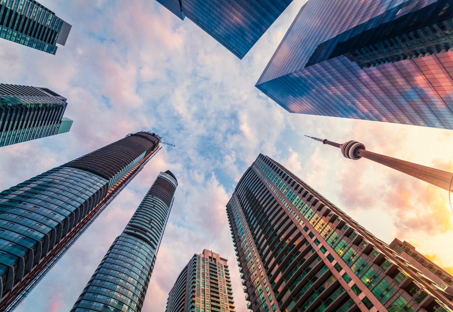 Upward view of modern skyscrapers and the CN Tower against a colorful sunset sky with soft pink and blue clouds.