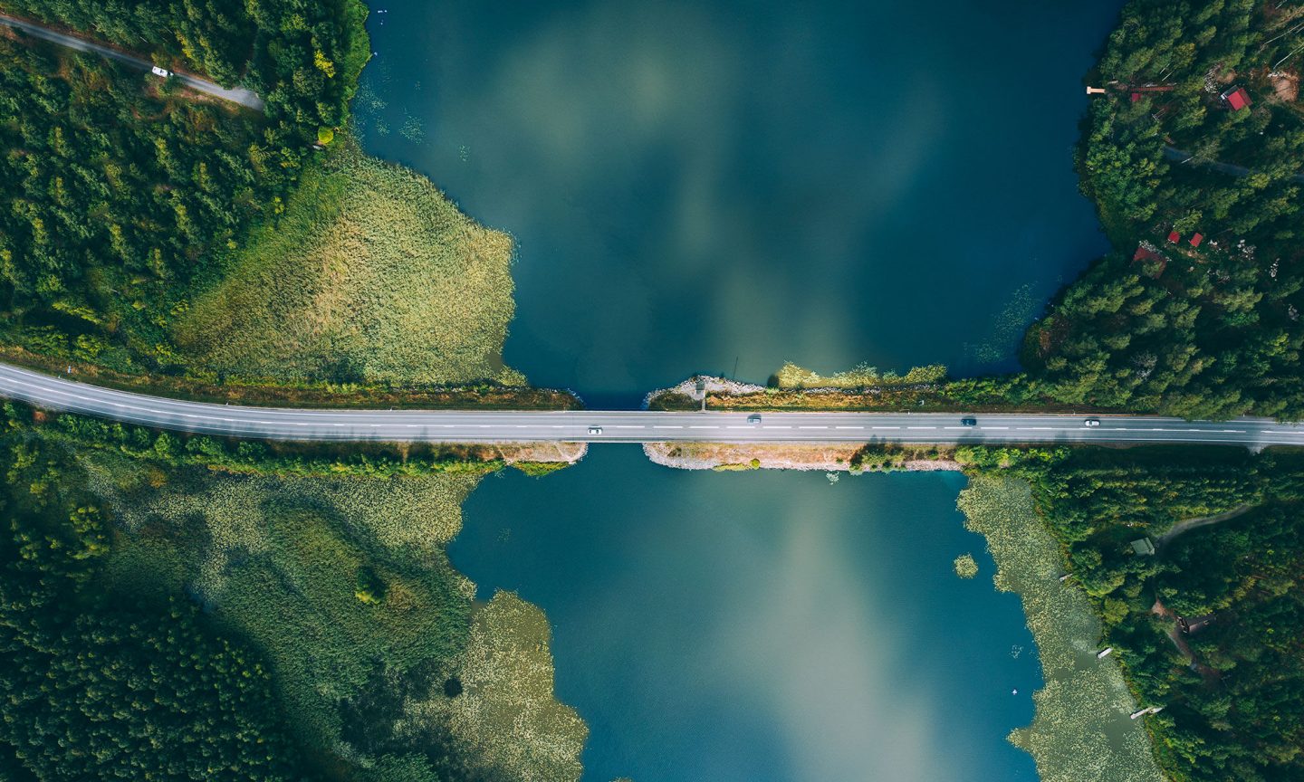 An aerial view shows a straight road bridge crossing a calm blue-green lake. Dense evergreen forest surrounds both sides of the water, creating a strong contrast between land and lake. The water reflects the sky, adding soft cloud patterns to the scene. The composition emphasizes symmetry and the isolation of the roadway cutting through the natural landscape.