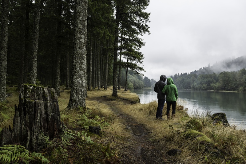 A couple wearing raincoats looks out over a foggy, misty lake in the fall in an evergreen forest in Washington state.