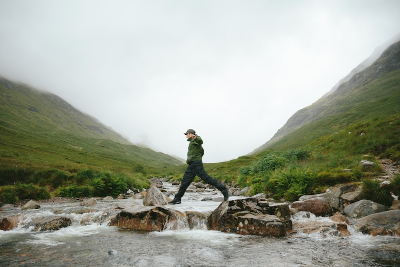man crossing a river over rocks in Scotland